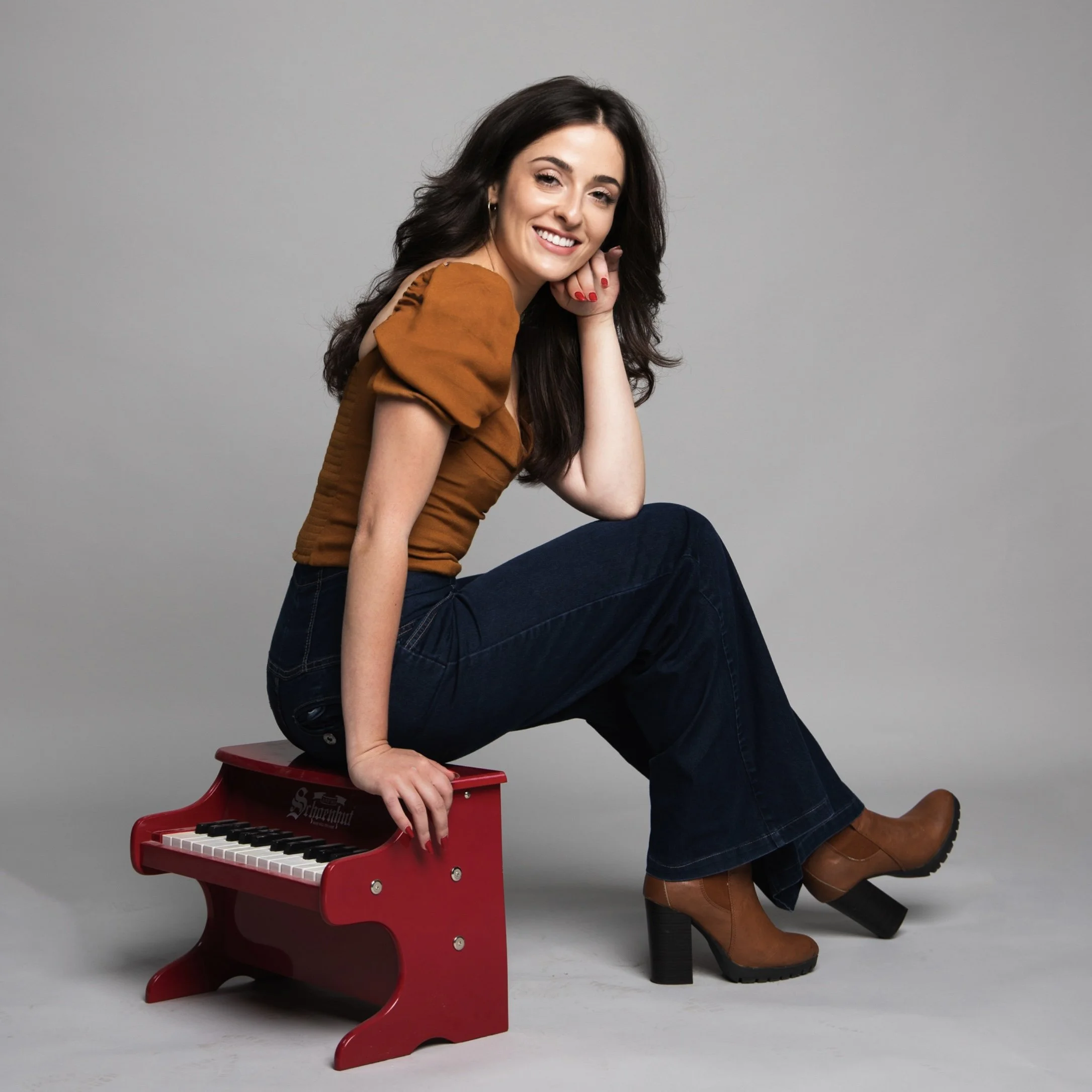A woman sitting on a small red toy piano, smiling and resting her head on her hand, wearing a rust-colored top, dark jeans, and brown heeled boots, in a studio with a gray background.